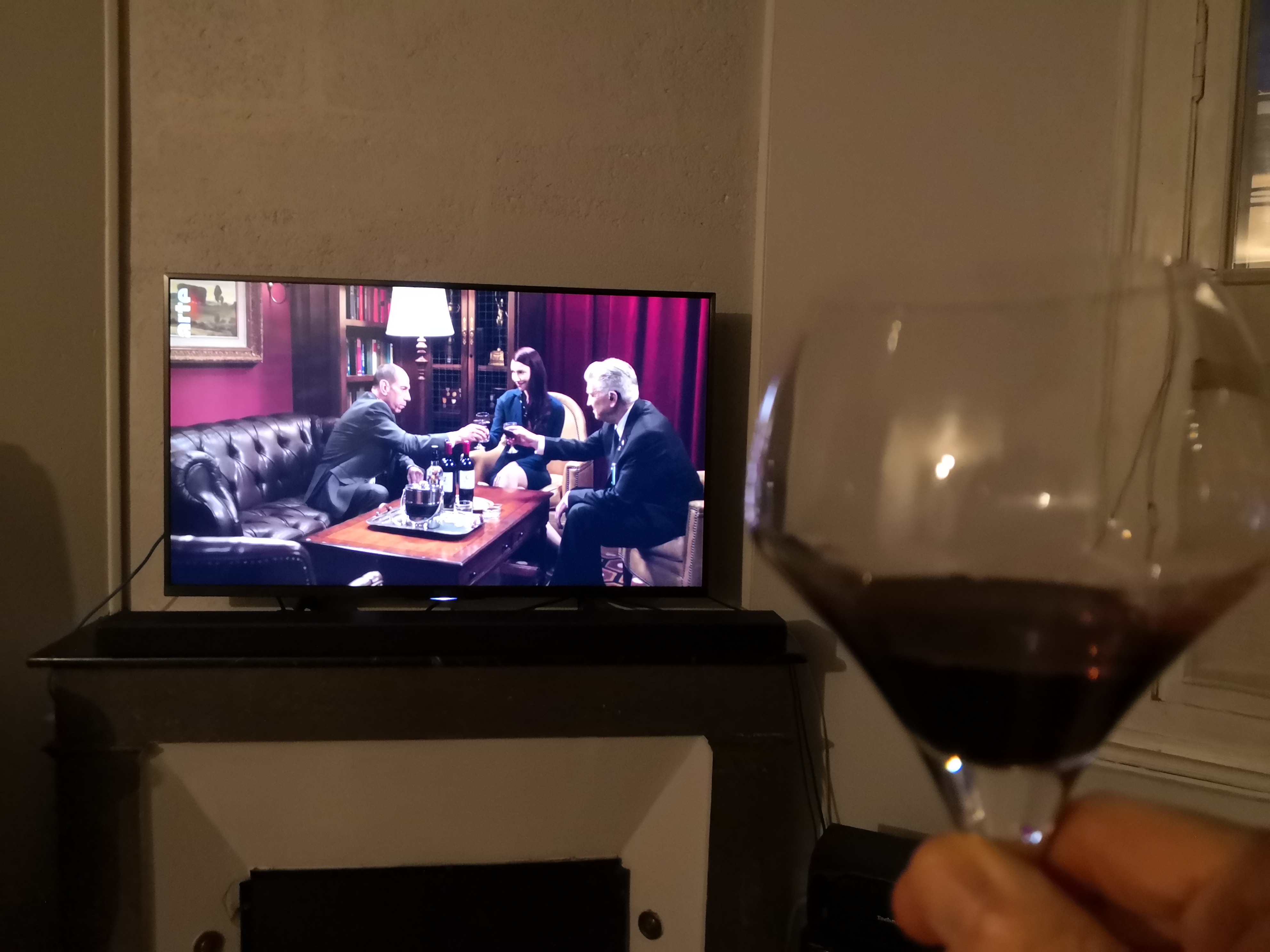 A hand raises a glass of red wine toward a television screen showing a scene from Twin Peaks in which Gordon Cole, Albert Rosenfield, and Tammy Preston are toasting her induction into the Blue Rose task force.